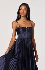A woman is wearing a navy blue pleated dress with spaghetti straps. The upper part of the dress has a ruched, satin-like fabric with a bow detail at the center, and the pleated skirt flows down gracefully. She is accessorized with long, dangling earrings.
