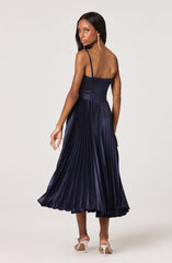 A woman is wearing a navy blue dress with thin spaghetti straps. The top section is fitted and ruched, and the skirt is high-waisted with a pleated design that falls to a midi length. She is also wearing white heels and long, dangling earrings.