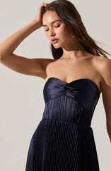 A woman is wearing a strapless, dark navy blue dress with a pleated texture. The top of the dress features a twisted knot detail at the center, creating a gathered effect. The dress is fitted around the bust with a smooth, shiny fabric, and the pleats extend from the waist down, giving the skirt a flowing appearance.