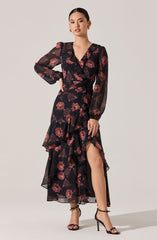 A woman is wearing a long, flowing dress with a black background and large, dark red and pink floral pattern. The dress features long, semi-transparent sleeves, a ruffled V-neckline, and a high slit on one side, revealing her leg. She pairs the dress with black high-heeled sandals with ankle straps.