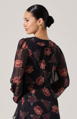 A woman is wearing a sheer black dress with long puffed sleeves, featuring a large floral pattern in shades of red and pink with greenish-brown leaves.