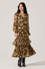 The dress is a long, brown floral print with gold and beige tones. It features a V-neckline, long puffed sleeves with elastic cuffs, and a tiered skirt with multiple ruffled layers. The fabric appears lightweight and slightly sheer, giving the dress a flowing, feminine silhouette. The woman is also wearing black knee-high boots.