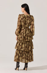 A woman is wearing a long, brown floral dress with a tiered skirt and puffed long sleeves. The dress features a gold and beige floral pattern throughout. She is also wearing black high-heeled ankle boots.