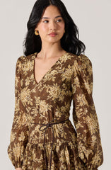 A woman is wearing a long-sleeved dress with a V-neckline and a floral print in shades of beige and cream on a brown background. The dress features puffed shoulders, a gathered waist with a thin tie, and a ruffled skirt.