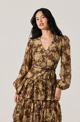 A woman is wearing a brown dress with a floral pattern of beige and light brown flowers and leaves. The dress has a V-neckline, long slightly puffed sleeves with elastic cuffs, a fitted waist, and a tiered skirt with ruffles.