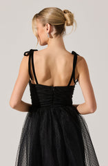 A woman is wearing a black dress with ruched fabric on the bodice, featuring thin shoulder straps that tie into bows. The skirt is made of a sheer, dotted tulle material. Her hair is styled in an elegant updo, and she is accessorized with sparkly, dangling earrings.