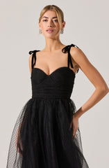 A woman is wearing a black dress with a fitted, textured bodice that has sweetheart neckline and shoulder straps tied into bows. The skirt of the dress is made of sheer, flowy material with a subtle dotted pattern. She is accessorized with large, rectangular earrings and has her hair styled in a sleek, middle-parted look.