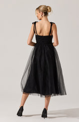 A woman is wearing a sleeveless black dress with shoulder straps tied into bows. The bodice is fitted with horizontal ruching, and the skirt is made of a sheer, polka-dotted tulle overlay that falls to mid-calf length. She has her hair styled in a low, messy bun and is wearing black high heels.