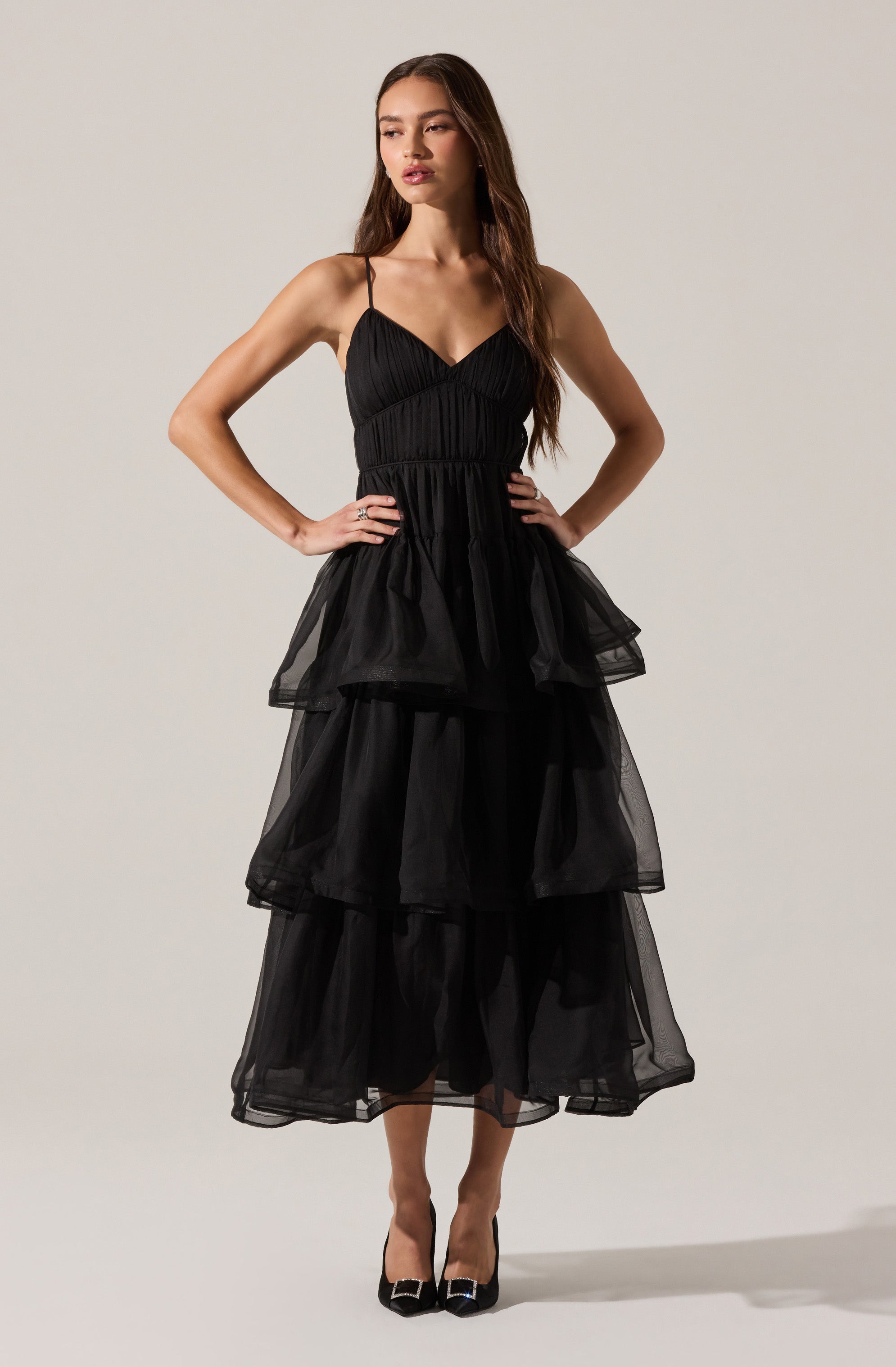 A woman is wearing a black dress with a fitted bodice and thin spaghetti straps. The dress features multiple tiers of voluminous, sheer fabric that create a layered and ruffled effect from the waist down to the hem. She pairs the dress with black high heels that have a pointed toe.