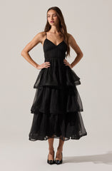 A woman is wearing a black dress with a fitted bodice and thin spaghetti straps. The dress features multiple tiers of voluminous, sheer fabric that create a layered and ruffled effect from the waist down to the hem. She pairs the dress with black high heels that have a pointed toe.