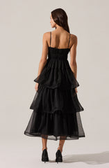 A woman is wearing a black sleeveless dress with thin spaghetti straps. The dress features a fitted, ruched bodice and a multi-layered, tiered skirt made of sheer, lightweight fabric that creates a voluminous, ruffled appearance. She is standing with her back to the camera, showing the back view of the dress.