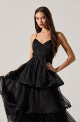 A woman is wearing a black dress with thin spaghetti straps, a fitted bodice with vertical pleats, and a layered, ruffled, and semi-sheer skirt made of lightweight fabric.