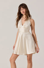 A woman is wearing a sleeveless, satin dress in a light cream color with a deep V-neckline. The bodice is fitted with gathered fabric at the shoulders, and the waist is accentuated with horizontal pleats. The skirt is flared and knee-length, made from shiny satin material. She accessorizes with a pair of gold earrings and a gold bracelet on her right wrist.