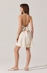 The woman is wearing a sleeveless, backless cream-colored dress with a high neckline, tied at the waist with a large bow. The skirt of the dress is short and flowy. She is also wearing cream-colored ballet flats with a small decorative bow on the toe.