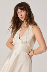 A woman is wearing a sleeveless, satin, off-white dress with a deep V neckline and a fitted waist. She has wavy, shoulder-length brown hair with bangs. She accessorizes with a gold bracelet on her right wrist and a matching gold hoop earring.