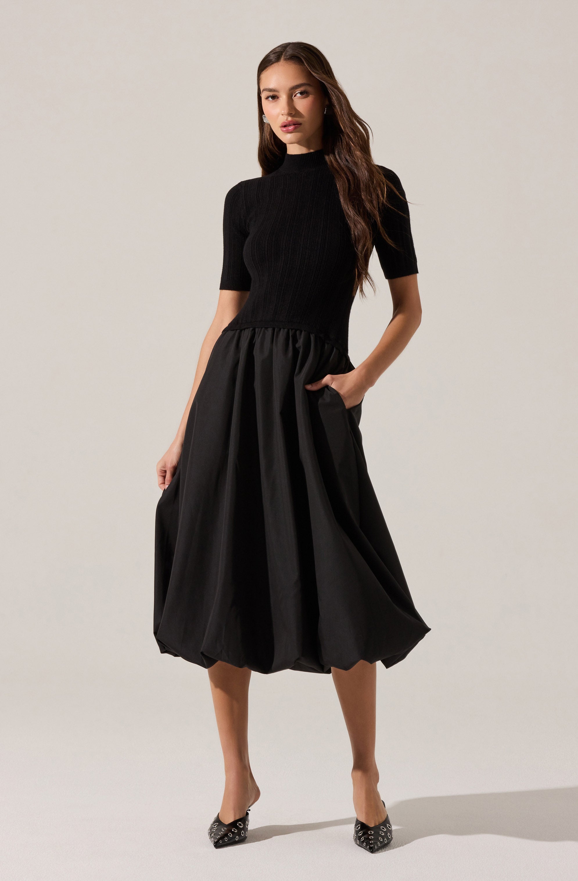 A woman is wearing a fitted black, short-sleeved, high-neck top with a ribbed texture, paired with a voluminous, knee-length black skirt that has a gathered, bubble-like silhouette. She is also wearing black high heels with paisley patterns.