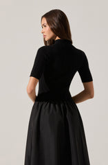 A woman is wearing a fitted, short-sleeved black ribbed sweater with a high neckline, paired with a high-waisted, full-length black skirt made of a shiny fabric that gathers at the waist.