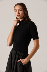 A woman is wearing a fitted, short-sleeved, ribbed black top with a high neckline. She pairs it with a black, high-waisted skirt that has a gathered waist and side pockets.