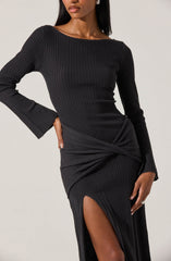 A woman is wearing a fitted, long-sleeved, ribbed black dress with a boat neckline. The dress features a gathered, draped section at the waist with a slit that reveals her leg. The fabric appears to be stretchy and textured.