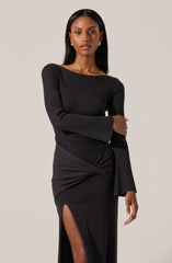A woman is wearing a fitted, long-sleeved, black ribbed knit dress with a boat neckline. The dress features a high slit on one side and a twist detail around the waist, creating a gathered effect.
