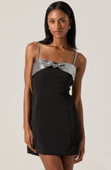 A woman is wearing a black dress with thin shoulder straps and a silver sequined overlay across the top, which forms a bow at the center.