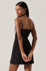 A woman is wearing a black sleeveless dress that features thin, adjustable, multicolored straps with metallic accents. The dress has a fitted bodice with a slight flare at the hem, creating a classic and elegant look.
