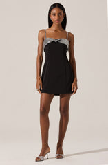 A woman is wearing a short, sleeveless black dress with thin, silver spaghetti straps. The dress features a silver-sequined panel across the chest area, and the hemline is above the knees. She is also wearing high-heeled shoes that are silver with a sparkling finish.