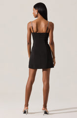 The dress is a black, fitted mini dress with thin, adjustable spaghetti straps. It features a sleek, minimalist design with a front zipper closure. The dress has a slightly A-line silhouette that flares out gently towards the hem. The model is wearing the dress with clear high-heeled shoes.