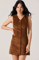 A woman is wearing a sleeveless, knee-length brown corduroy dress with a V-neckline. The dress features a row of fabric-covered buttons down the front and has two flap pockets at the waist. She is accessorized with gold earrings, a gold bracelet, and a ring.