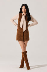 A woman is wearing a beige long-sleeve shirt with a large beige scarf tied around her neck, a brown sleeveless button-down dress that reaches mid-thigh, and matching knee-high brown suede boots.