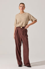 A woman is wearing a beige, short-sleeved, slightly oversized knit sweater with a round neckline. She pairs it with high-waisted, wide-leg, rust-colored trousers that have pleats at the waist and a belt. She is also wearing black shoes and gold earrings.