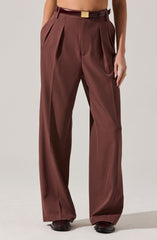 The image shows a person wearing high-waisted, wide-leg brown trousers with pleats at the waist. They have belt loops and are accessorized with a dark brown belt featuring a gold rectangular buckle. The trousers are tailored with creases running down the front of each leg.