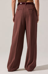 The image shows a person wearing high-waisted, wide-leg trousers in a rich brown color. The trousers have a tailored fit with pleats at the front, and feature two back welt pockets with button closures. The person is also wearing a dark brown belt, which complements the trousers. The overall style appears to be formal or business casual.