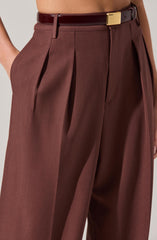A person is wearing high-waisted, wide-leg brown trousers with pleats at the waist. The trousers are paired with a dark brown belt with a large gold buckle. The fabric appears to be a smooth, slightly shiny material.