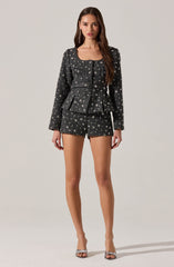 A woman is wearing a matching black and silver sequined blazer and shorts set. The blazer has long sleeves and a round neckline, with a front opening that features buttons. The shorts are high-waisted, fitted, and also adorned with a silver star pattern matching the blazer. She is wearing open-toed high-heeled sandals with a glittery finish.