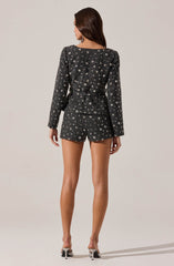 A woman is wearing a matching set consisting of a long-sleeved blazer and shorts. The blazer is black with a pattern of white stars and appears to be made of a textured fabric, with a tailored fit and a belt at the waist. The shorts match the blazer in pattern and texture, providing a coordinated look. She is also wearing high-heeled shoes with thin heels.