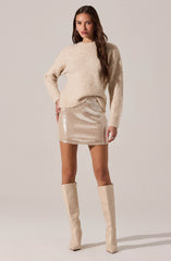 A woman is wearing a cozy, oversized, cream-colored knit sweater with long sleeves. She pairs it with a short, shimmering, champagne-colored sequin skirt that falls mid-thigh. She completes the look with knee-high, glossy, beige boots.