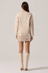 A person is wearing a loose, cream-colored knitted sweater with long sleeves and a round neckline. They are also wearing a short, shiny, beige mini skirt that appears to be made of a sequined or metallic fabric. The outfit is completed with knee-high, beige stiletto boots.