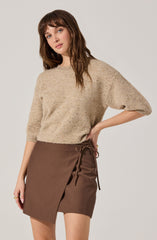 A woman is wearing a beige, speckled, slightly loose-knit sweater with three-quarter length sleeves. She pairs it with a brown wrap skirt that has a side tie closure.