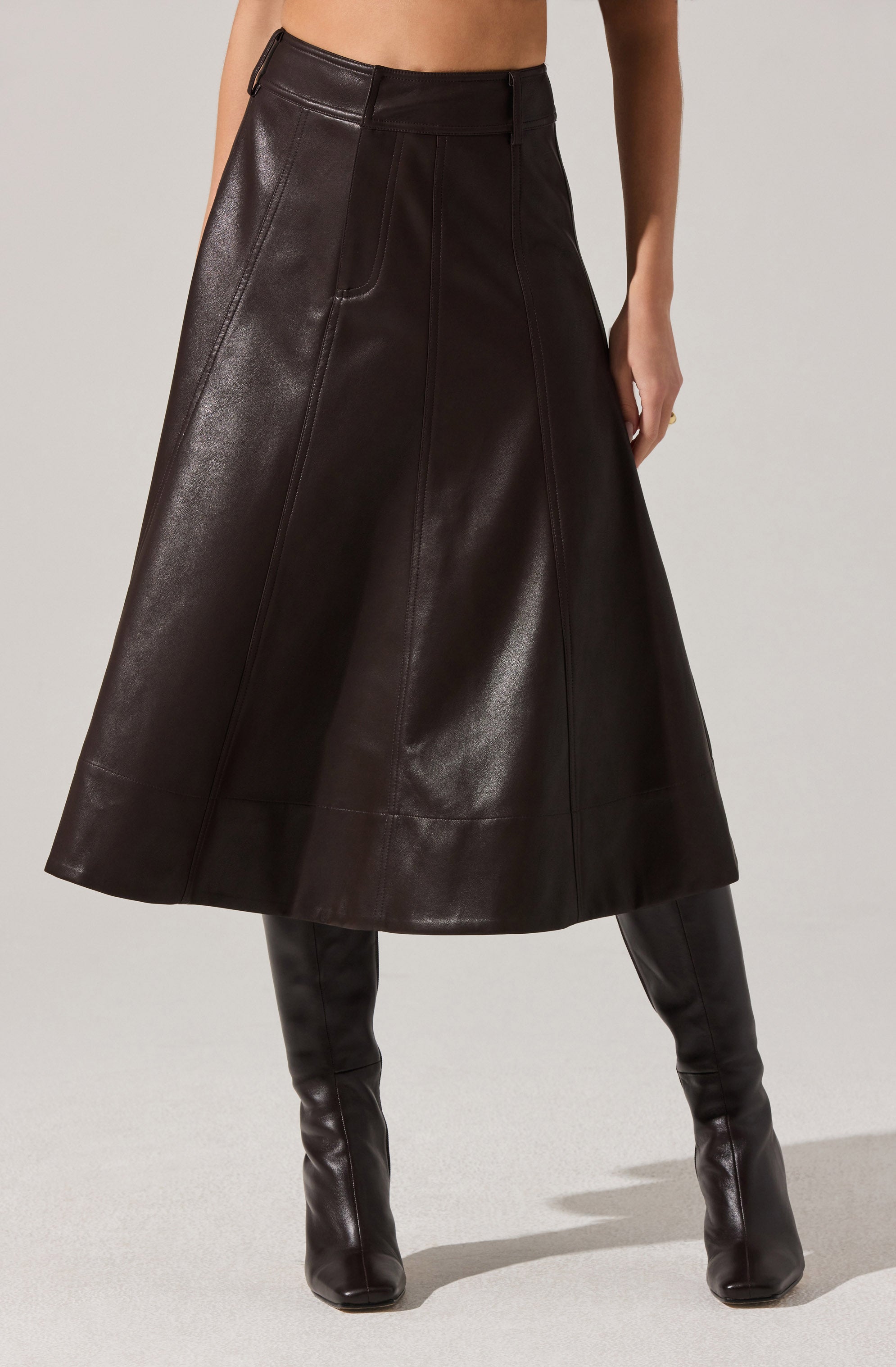 A person is wearing a long, dark brown leather skirt that reaches mid-calf. The skirt features vertical seam details and a high waist with belt loops. They are also wearing knee-high black leather boots. The person's top is not visible in the image.