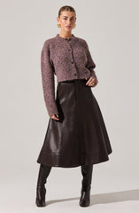 A woman is wearing a textured, long-sleeve, button-up sweater in shades of purple and pink, paired with a high-waisted, A-line black leather skirt that reaches below her knees. She completes the look with black knee-high leather boots and large, gold hoop earrings.
