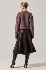 A woman is wearing a textured, multicolored, cropped sweater in shades of purple and pink, paired with a high-waisted, knee-length black leather skirt that has a flared silhouette. She is also wearing tall black leather boots with stiletto heels.