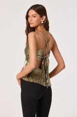A woman is wearing a sleeveless, metallic gold top with thin straps that cross at the back and tie in a bow. The top has a pleated design and features a small keyhole opening in the back. She pairs it with black pants.