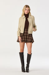 A woman is wearing a beige cropped jacket with a collar and elastic cuffs, a dark brown turtleneck top, a high-waisted plaid brown skirt, and tall black leather boots.