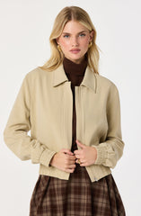 A woman is wearing a beige zip-up jacket with elasticized cuffs and collar, over a dark brown turtleneck top. She is also wearing a brown plaid skirt. She has blonde hair styled in loose waves and is accessorized with gold hoop earrings and a gold ring.