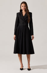 A woman is wearing a black tailored blazer with a cropped fit and lapels, paired with a high-waisted, full-length black skirt that has pleats. She is also wearing black pointed-toe shoes with a bow detail.