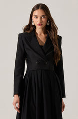 A woman is wearing a black outfit that features a fitted blazer with a wide lapel collar and two buttons at the front, paired with a long, pleated skirt. The blazer has a tailored fit with structured shoulders, and the skirt has a flowing, gathered design. She is accessorized with large, statement pearl earrings.