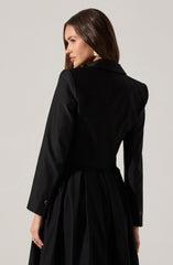 A woman is wearing a black tailored jacket with a structured fit and a notched collar. The jacket has long sleeves with buttoned cuffs. She is also dressed in a matching black pleated skirt that extends below the waist. Her accessories include large, round earrings.