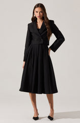 The outfit consists of a black cropped blazer with a tailored fit and double-breasted buttons, featuring a lapel collar and long sleeves. The skirt is a high-waisted, full-length, pleated design made from a medium-weight fabric, creating a flowing silhouette. The model is also wearing black pointed-toe shoes with a decorative bow detail on the front.
