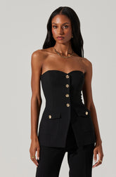 A woman is wearing a strapless black blazer with gold buttons down the front and on the pockets. The blazer has a fitted silhouette and two flap pockets, each with a gold button. She pairs it with matching black pants. She is accessorized with a gold chain necklace and gold earrings.